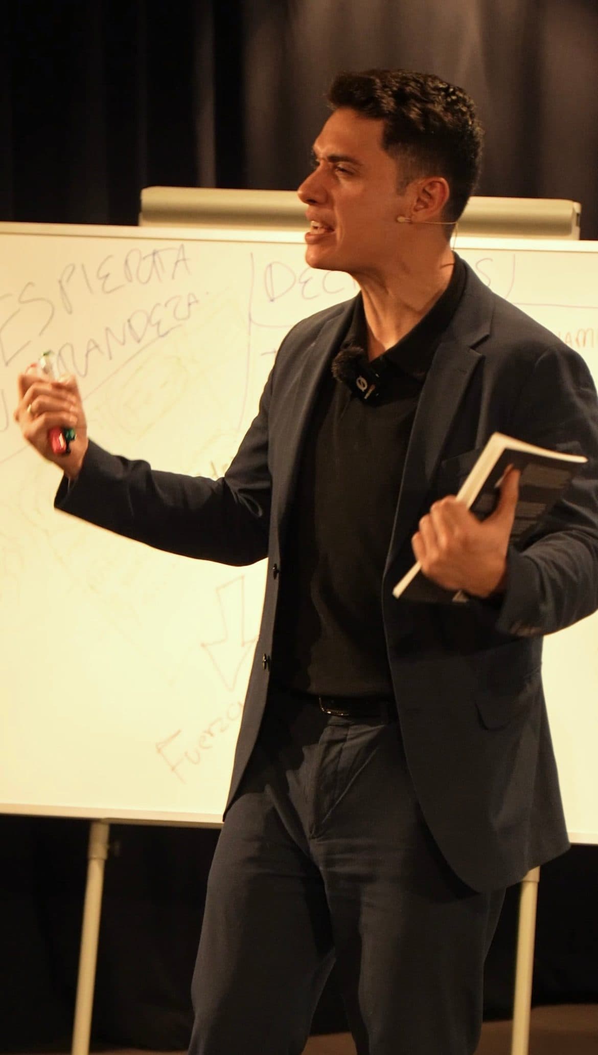 Carlos Medina — conferencia editorial, libro en mano frente a pizarra